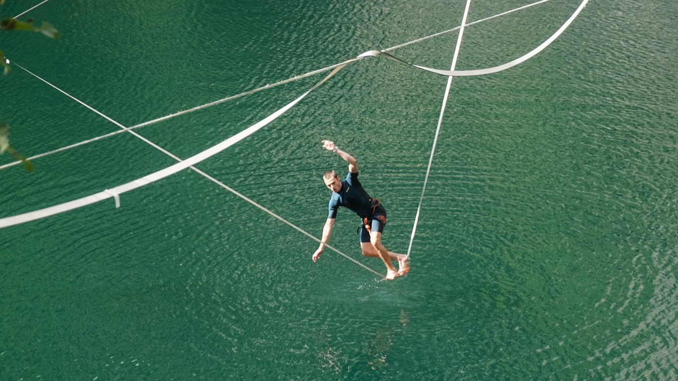 Slackliner balanciert über einem türkisgrünen See auf einer Slackline und hält das Gleichgewicht mit ausgestrecktem Arm.